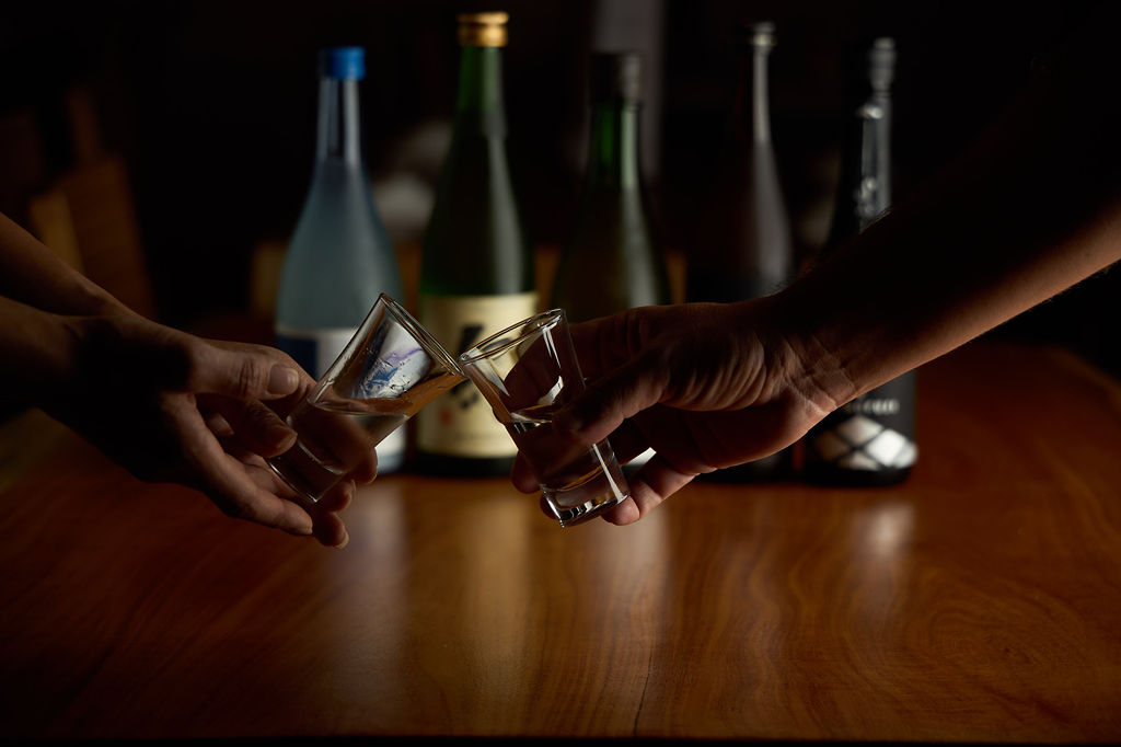 Two guests toasting with sake glasses at Anaba, bottles of premium sake in the background